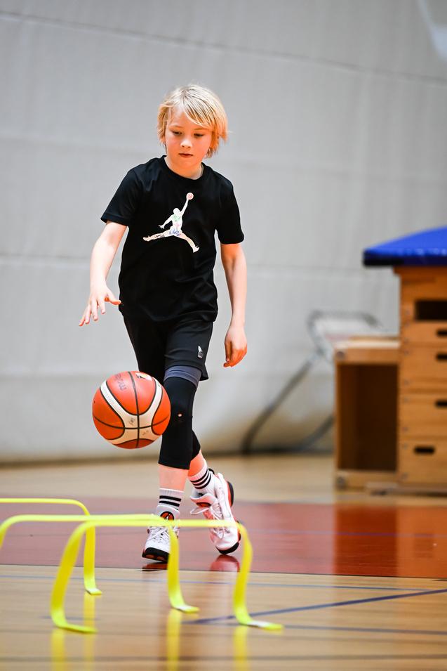 Junge dribbelt einen Basketball über gelbe Hürden in einer Sporthalle. Er trägt ein schwarzes T-Shirt und Sporthose.
