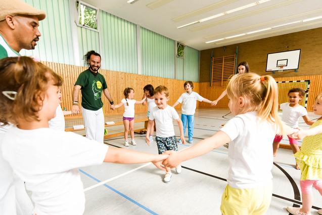 Gruppe von Kindern in T-Shirts und Erwachsenen in einer Turnhalle, die gemeinsam einen Kreis bilden und spielen.
