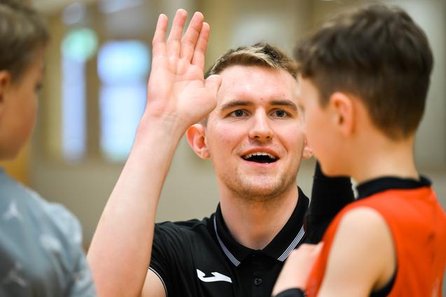 Trainer spricht mit einem jungen Spieler, während er seine Hand hebt, in einer Sporthalle mit Holzwand.